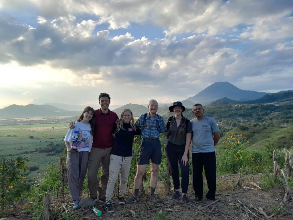 Six individuals pose for picture on a hill with mountains and farmland in the background.