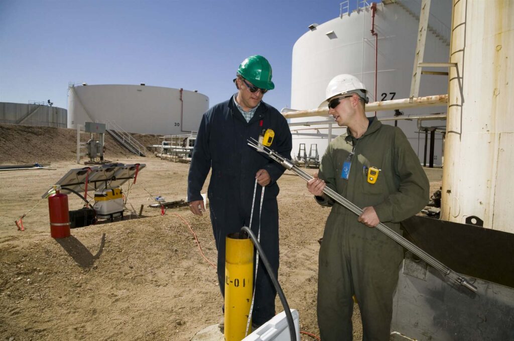 Two men in hardhats stand outside at an oil refinery site.