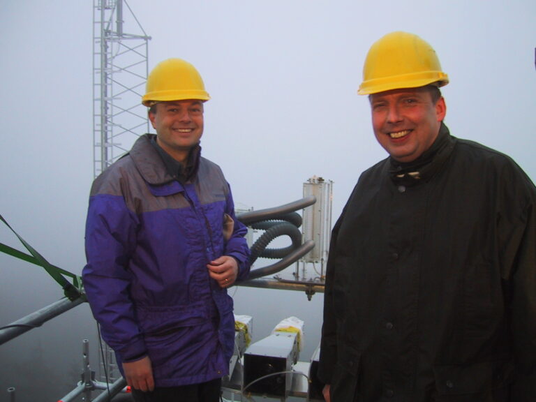 Collett and another researcher, both in hard hats, pose for an informal portrait in a foggy setting with tower structure visible behind them, and land dimly visible below.