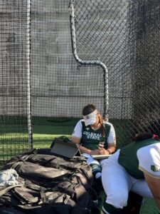 Woman in softball uniform sits in the shade on a pile of bat bags, studying.