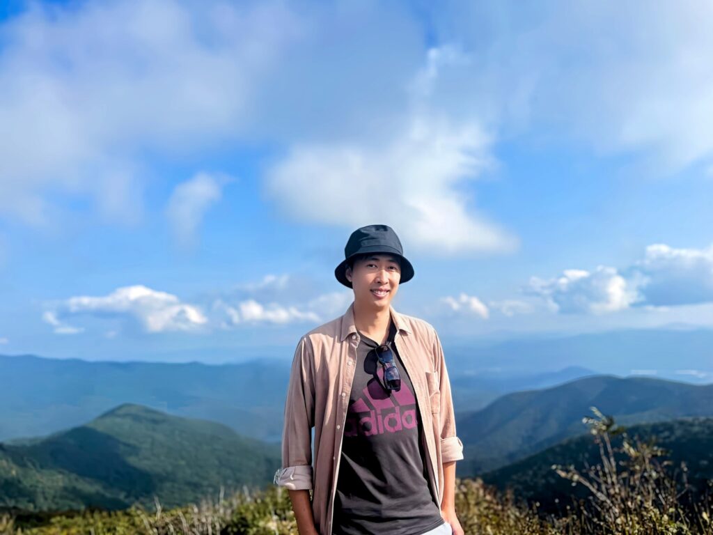 Casual outdoor portrait of a man in a hat posing in front of a mountainous landscape