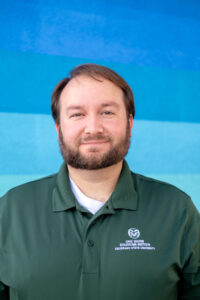 A posed portrait photo of Tyler Wible. He is wearing a green polo shirt with a CSU Ram's Head logo and the words "ONE WATER SOLUTIONS INSTITUTE COLORADO STATE UNIVERSITY" embroidered on it.