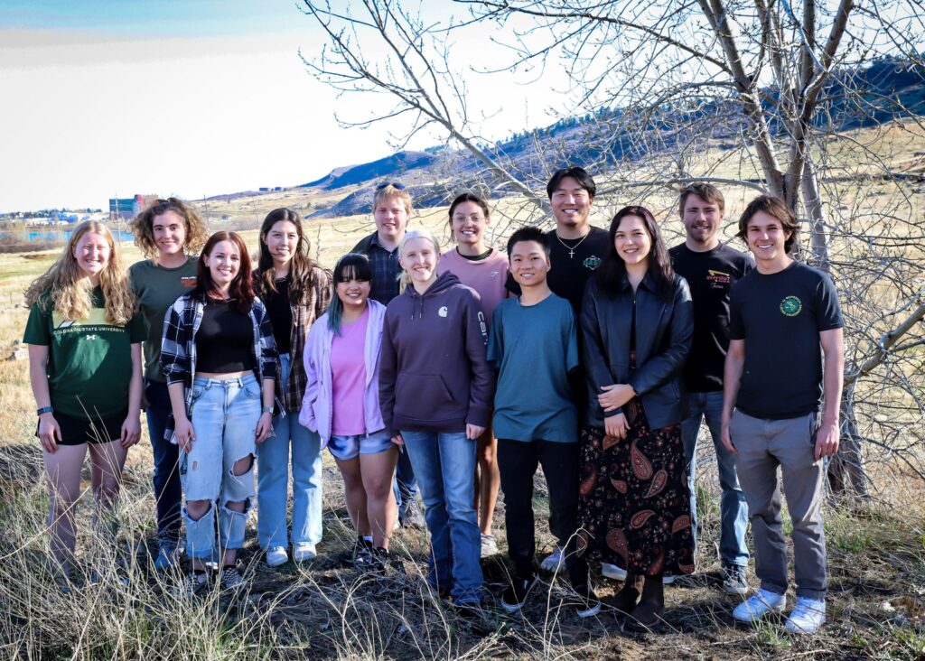 A large group of BMES students pose for a casual outdoor portrait in the foothills with Jian Cohen, a fellow student born without arms.