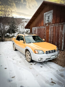 A yellow Subaru Baja with grey bumpers and fender flares sits on a snowy gravel driveway in front of a red barn.