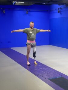 An adult male stands with his arms in a “T” in a gait lab. He is standing on a strip in the middle of the room’s floor which is equipped with sensor pads. The room has bright blue walls and multiple cameras spaced around the perimeter trained on the subject.