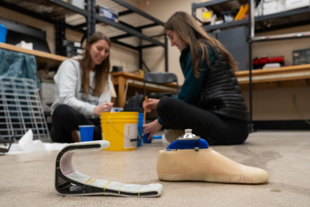 In the background, two female students sit on the floor surrounded by materials. They are mixing materials in a cup with wooden sticks. In the foreground sits two prosthetic feet. The one of the left is basic and simplistic, and made out of black/white plastic-like material. The one on the right resembles more of a typical human foot.